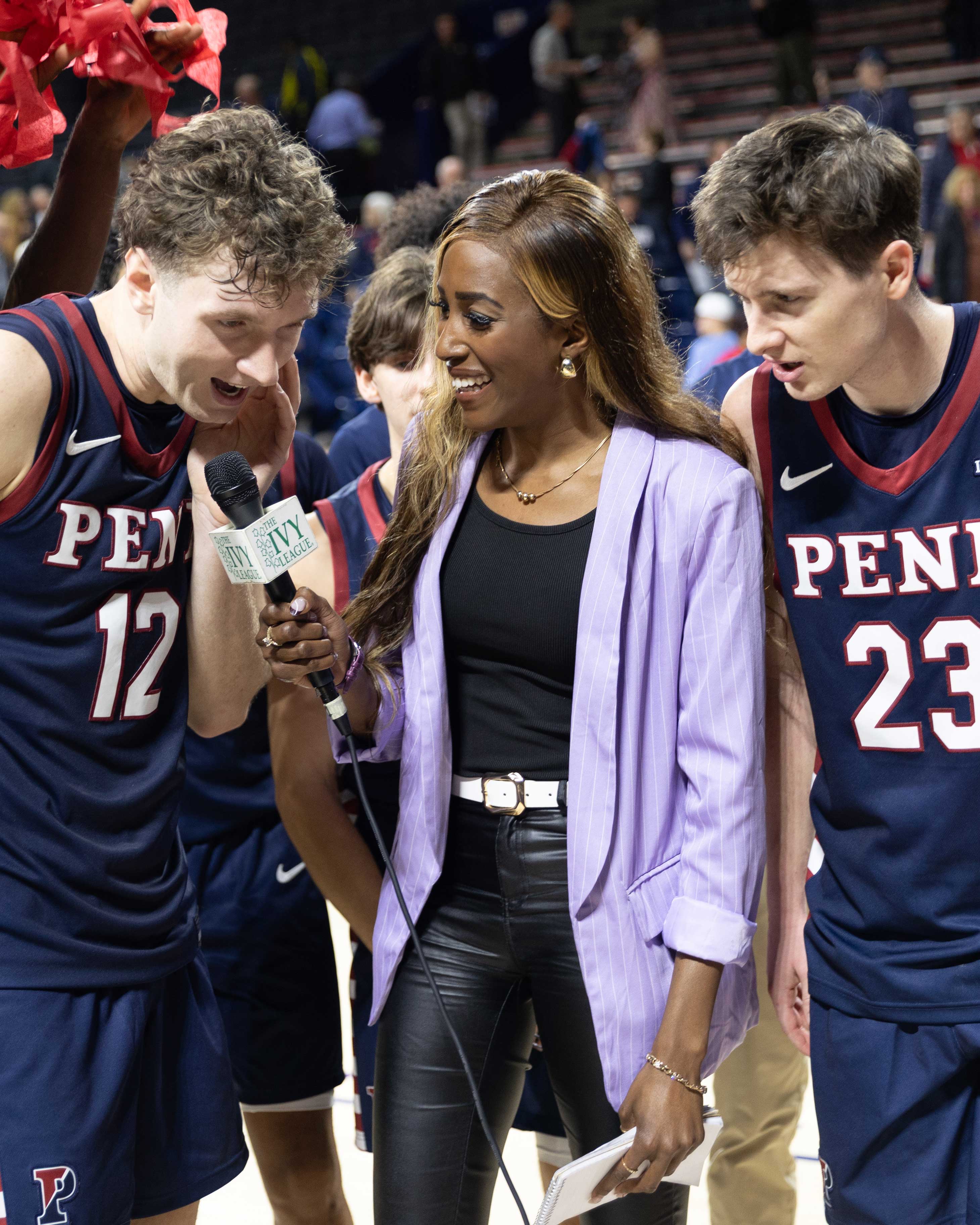 Renee wears a light purple blazer and black leather pants; flanking her are two basketball players mid interview.