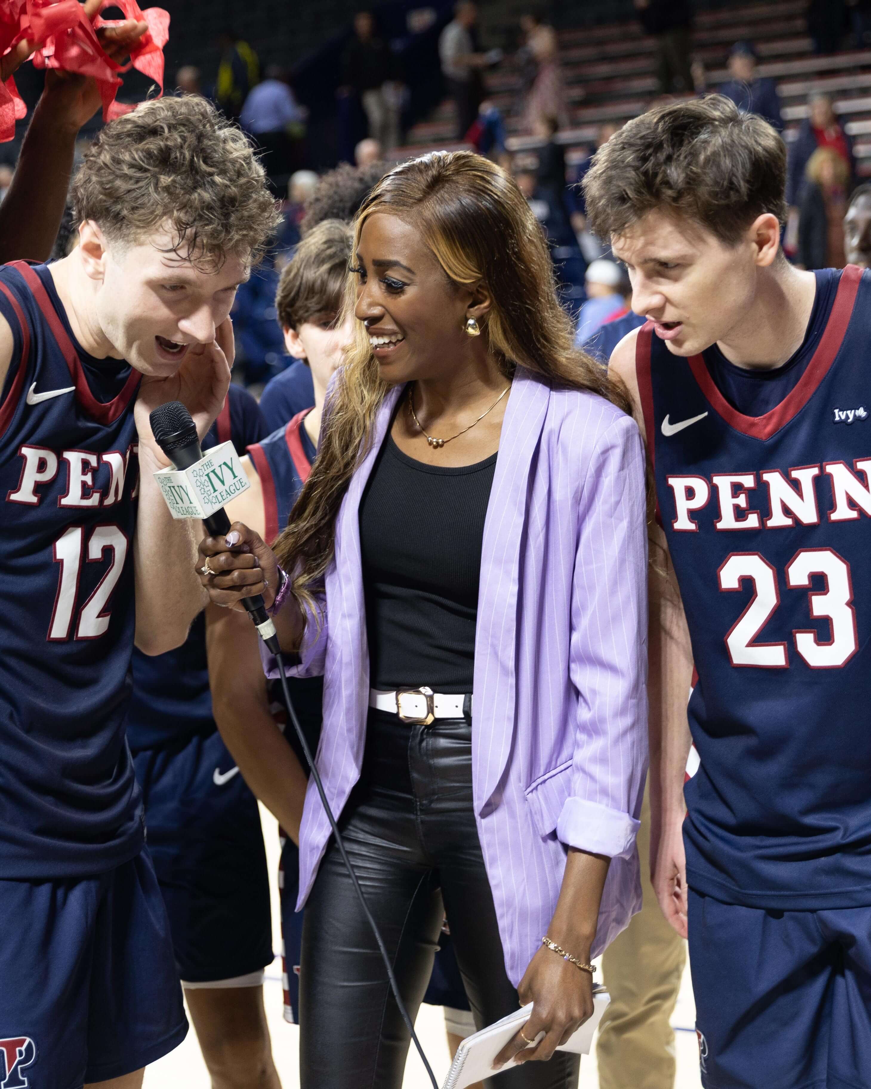 Renee wears a light purple blazer and black leather pants; flanking her are two basketball players mid interview. 