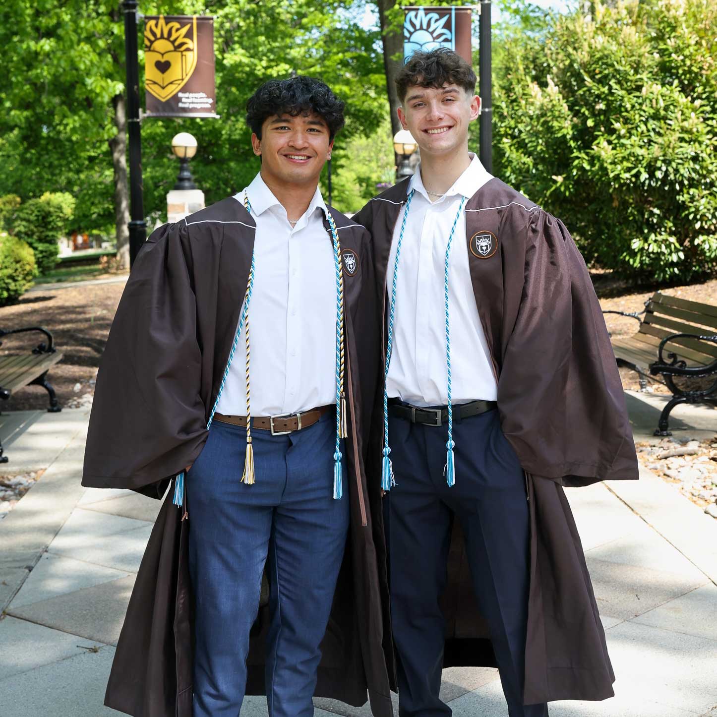 Sabato and Andrei wear their graduation robes over white shirts and blue slacks. They smile and pose on campus.