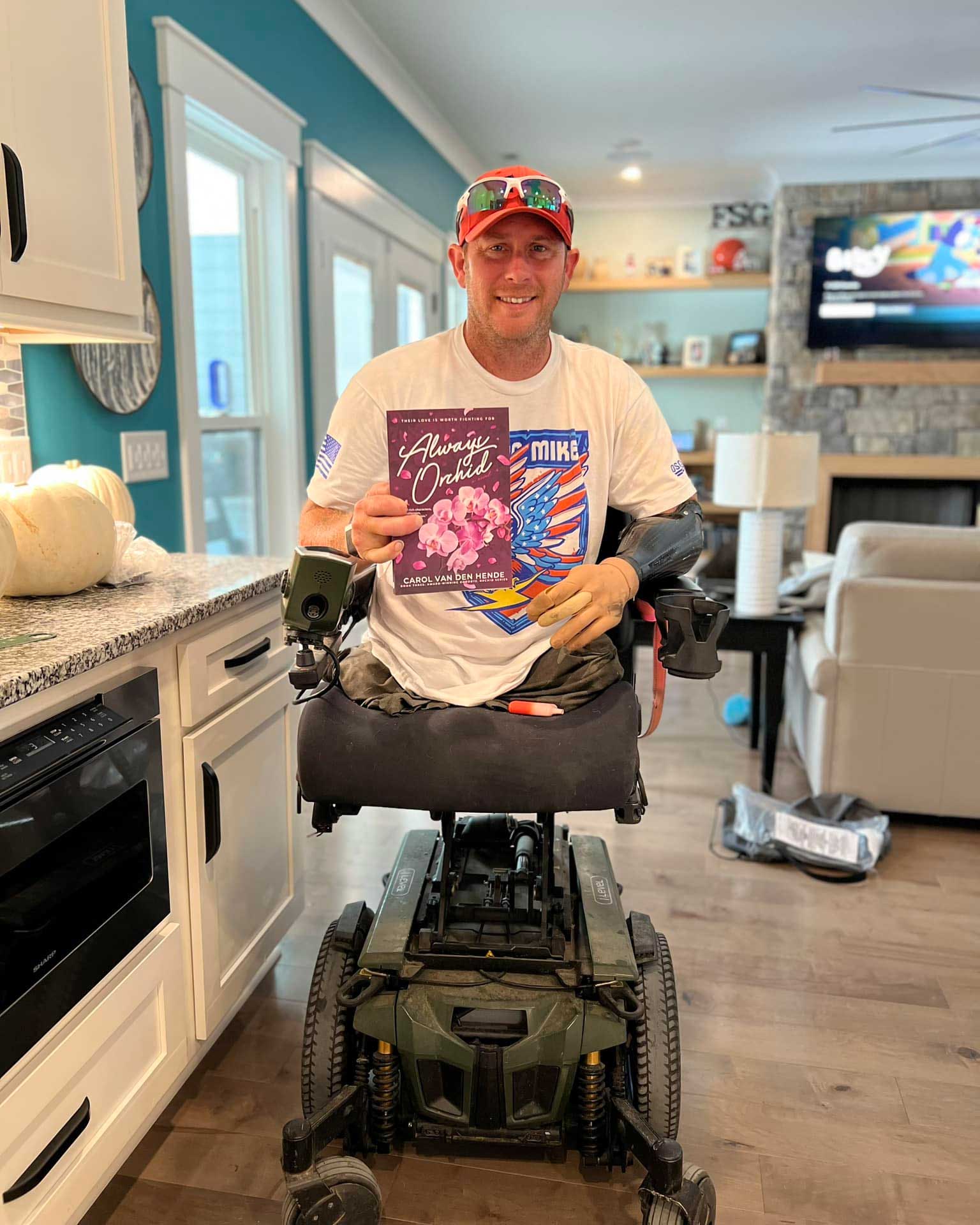 Sgt. Bryan Anderson holds up Carol's book 'Always Orchid' and smiles. He sits in a motorized wheelchair and wears a white t-shirt.