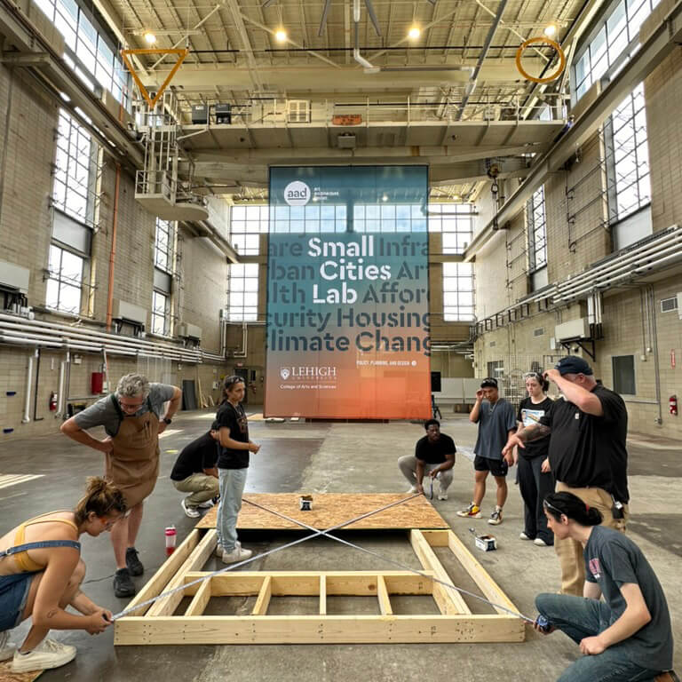 A group of people assembles a wooden structure inside a large industrial building with a banner about urban infrastructure and climate change in the background.