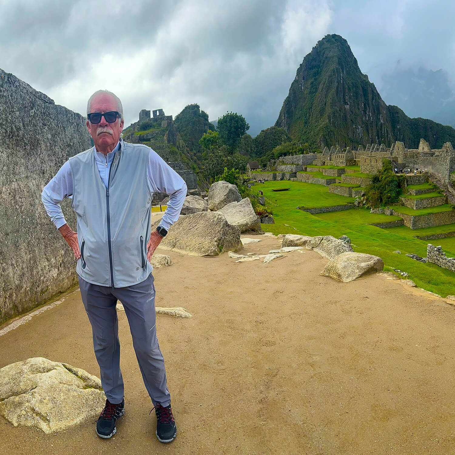 A man stands at Macchu Picchu with stone ruins and a mountain in the background on a cloudy day.