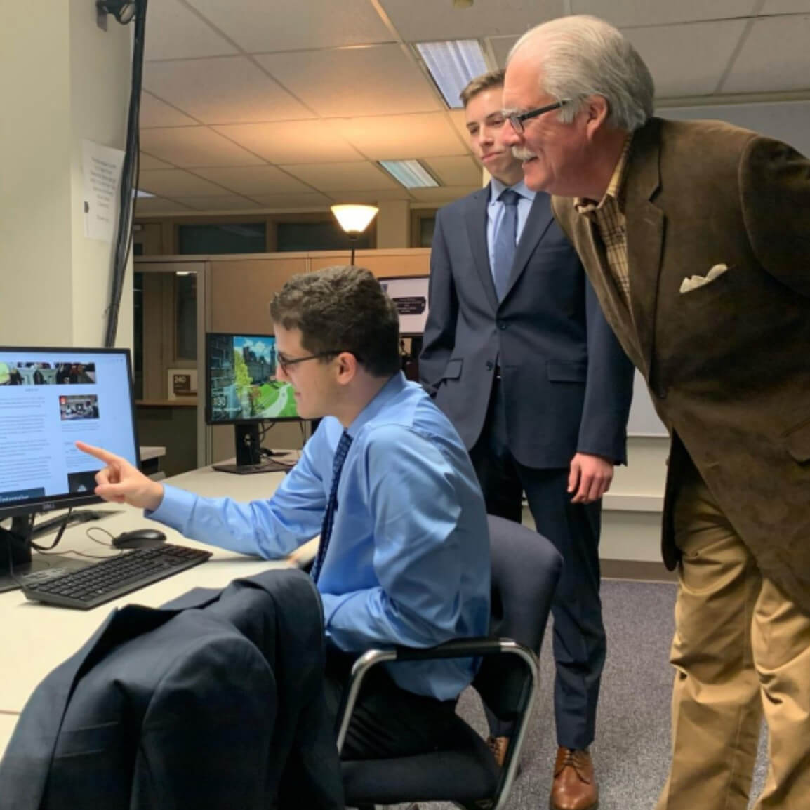 Three men in business attire gather around a desk in an office, with one seated and pointing at a computer screen while the two others are leaning in and looking at the display.