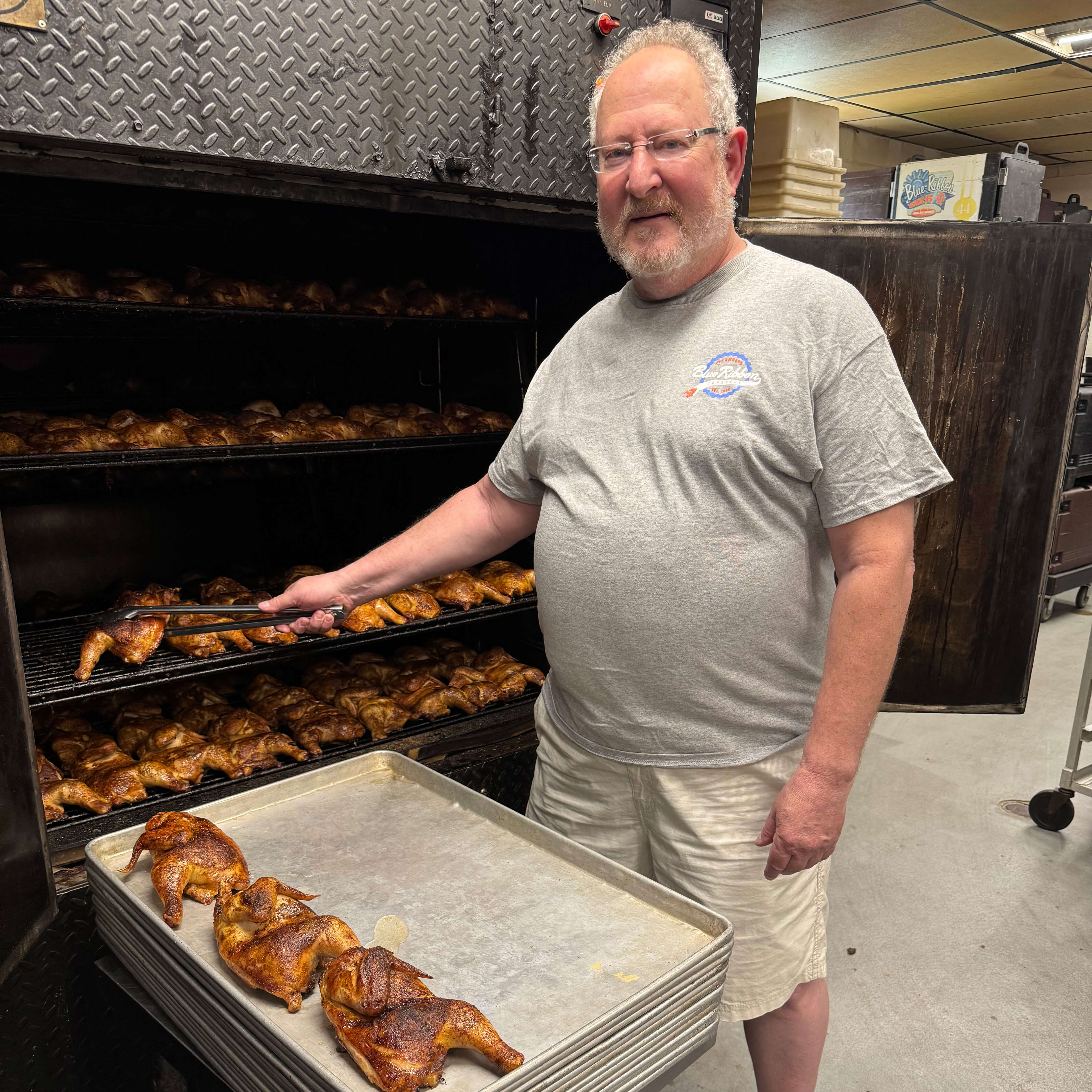Man in a gray T-shirt holding tongs, pulling roasted chicken from a large smoker.