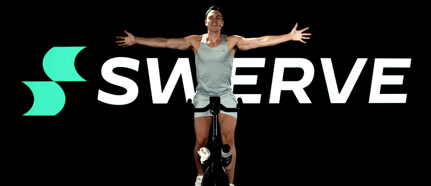 A man on a spin bike sits up on the bike with his arms outstretched in front of a sign that reads "Swerve".