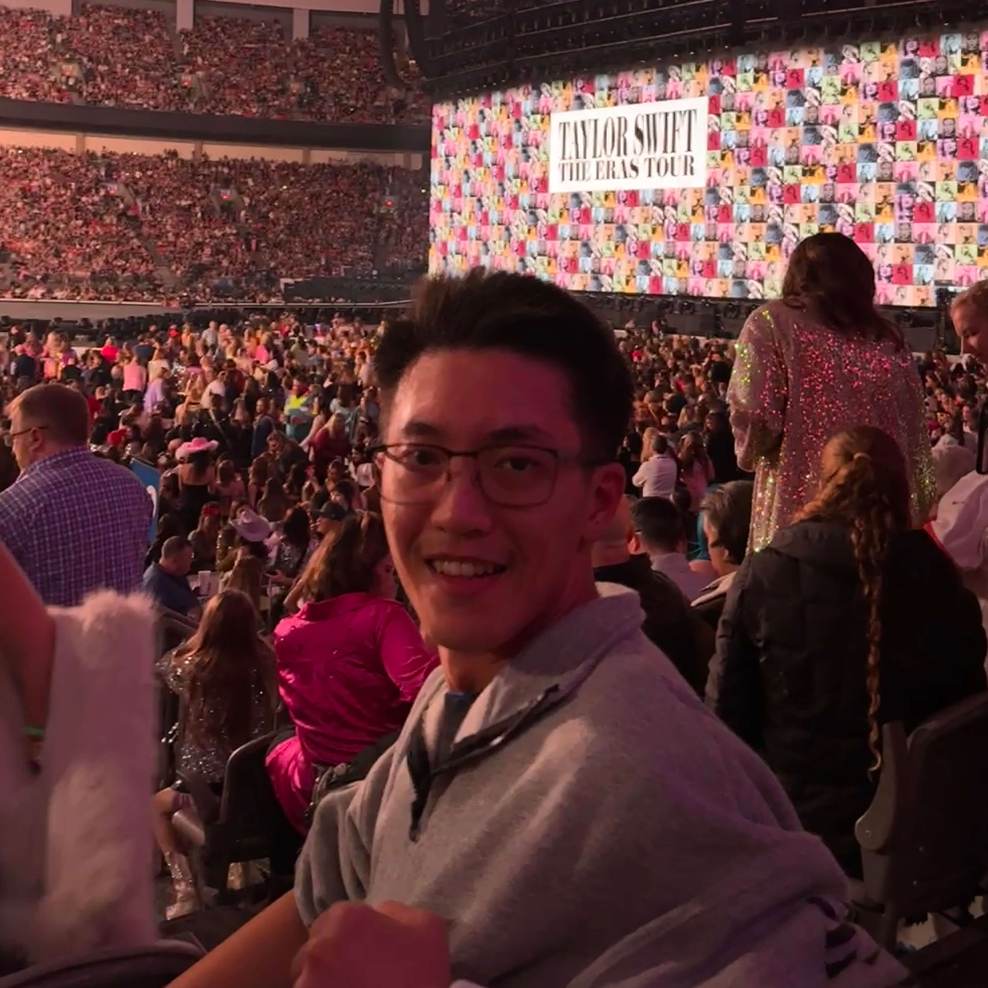 Winton smiles and turns to face the camera. Behind him are swaths of people and the multi-colored Taylor Swift Eras Tour logo. 