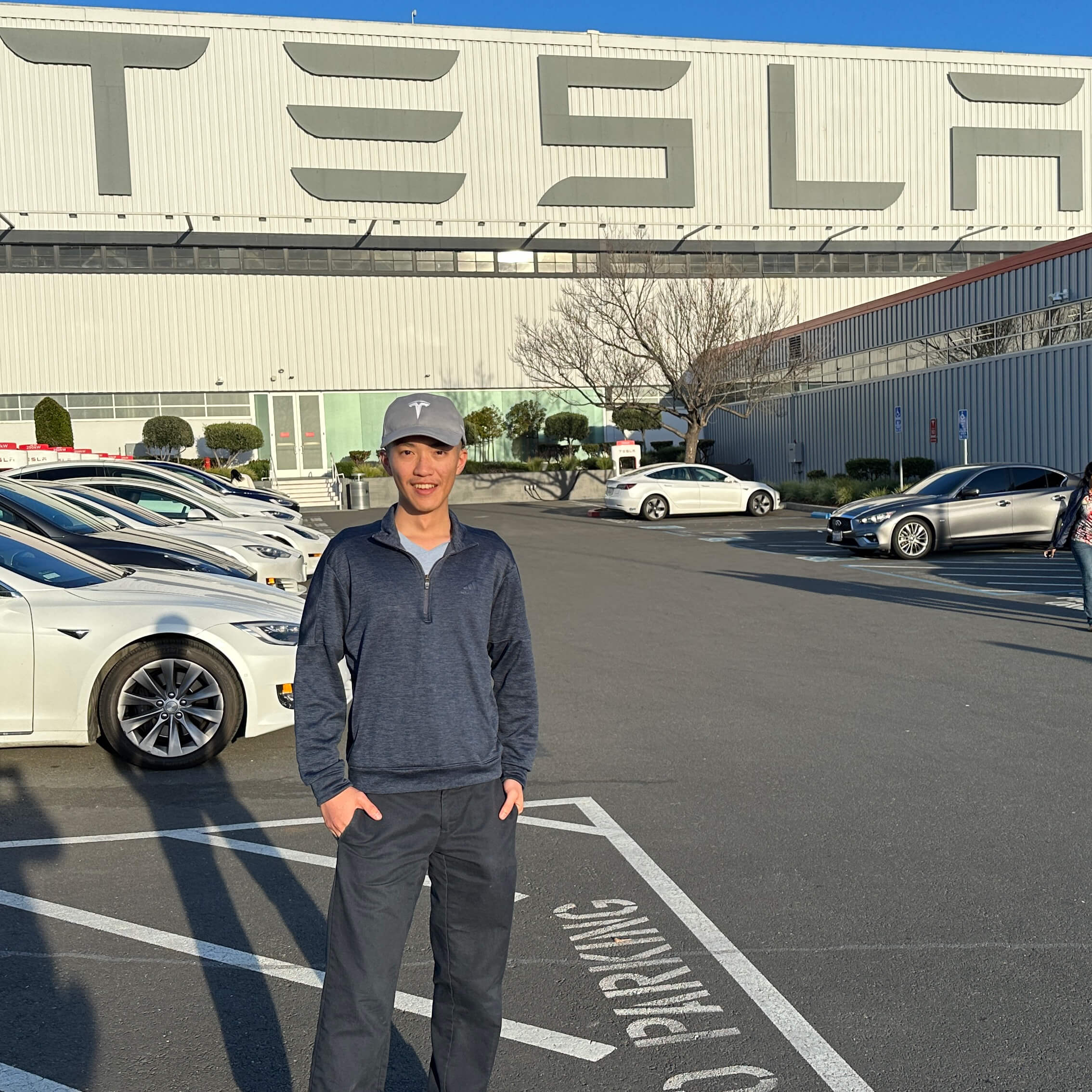 Winton smiles and stands in a Tesla parking lot in front of the facility. He wears a tesla-branded hat and blue quarter zip.