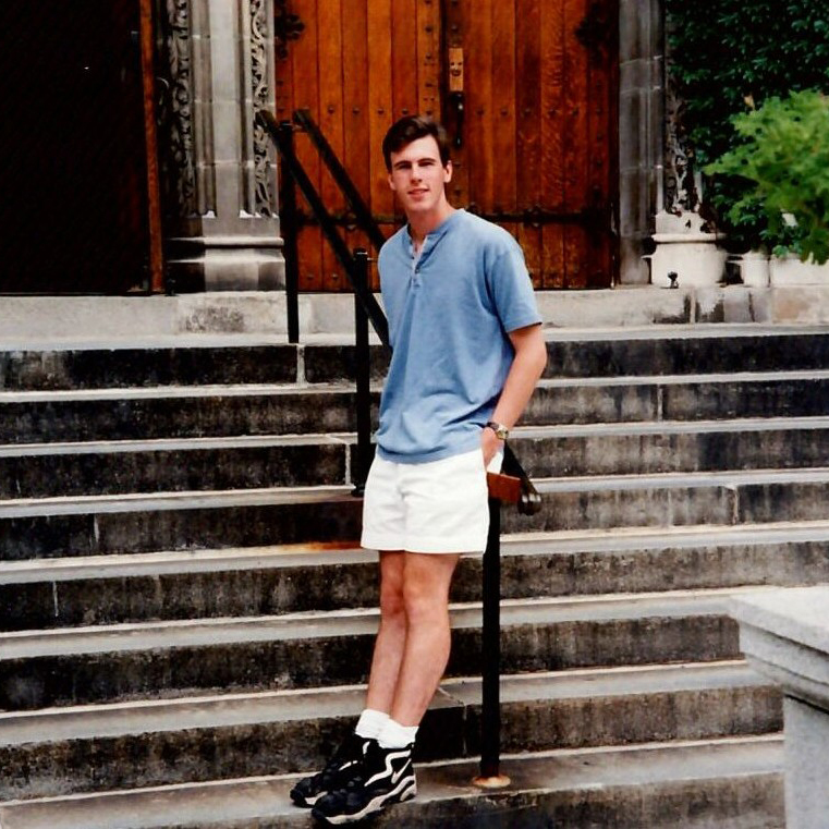 A young Brian leans against the railing of the steps leading to Packer Memorial Church. He wears a blue polo and white shorts.