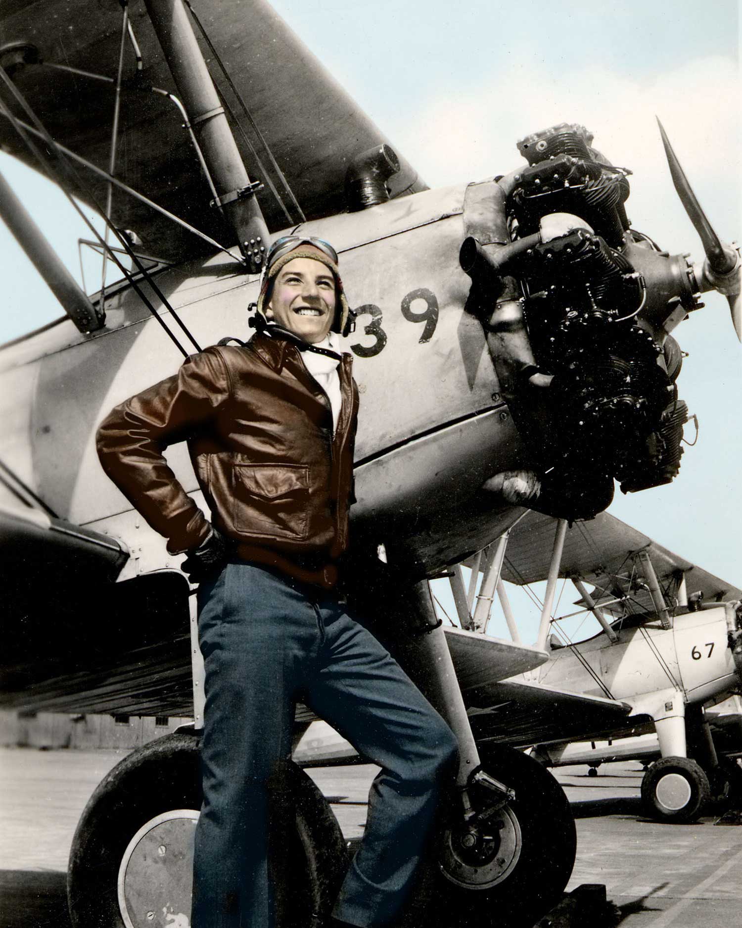 Alan stands in front of an World War II aircraft and smiles. He wears an aviator hat, goggles, and a dark brown leather jacket.
