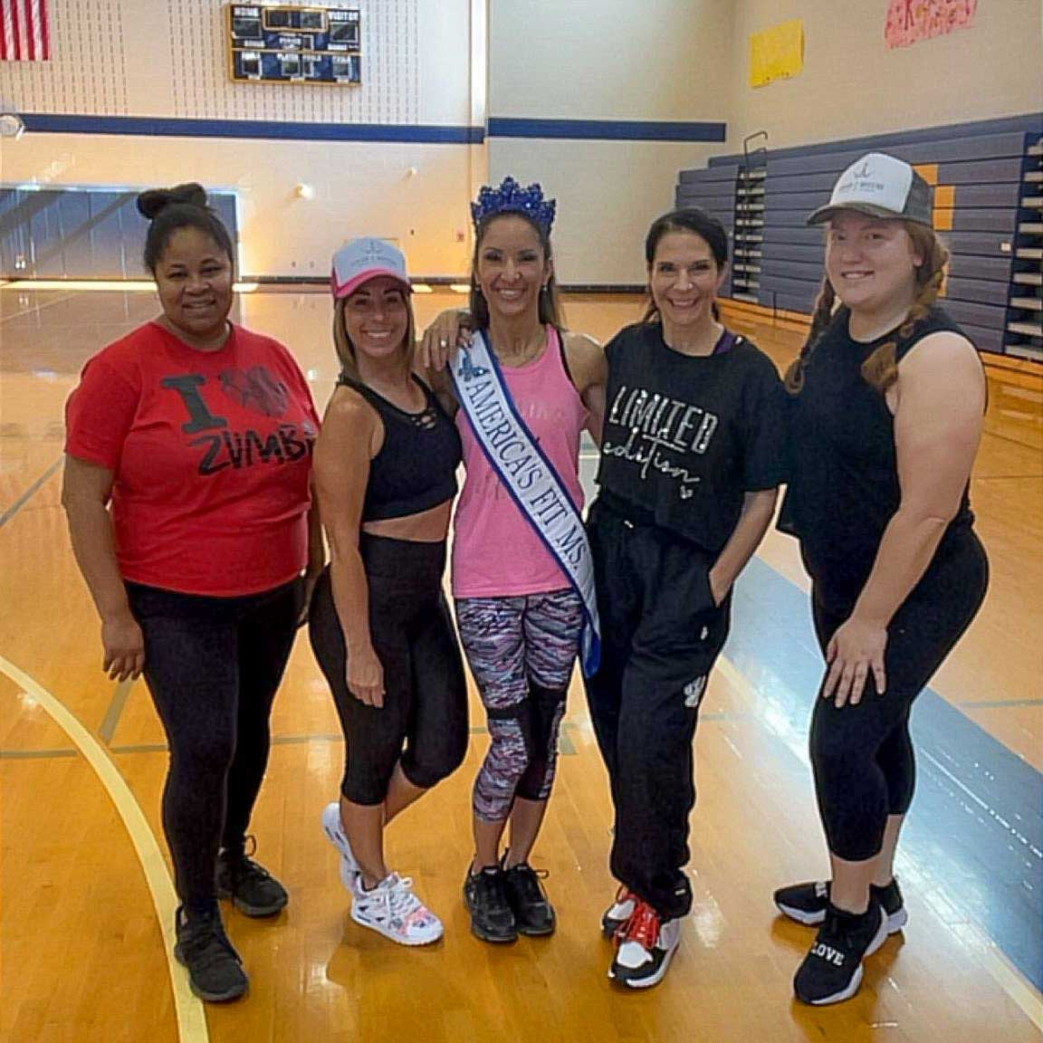 Kate poses with four other Zumba instructors in Broughal's gym. Kate wears a crown and a sash that reads 'America's Fit Ms.'