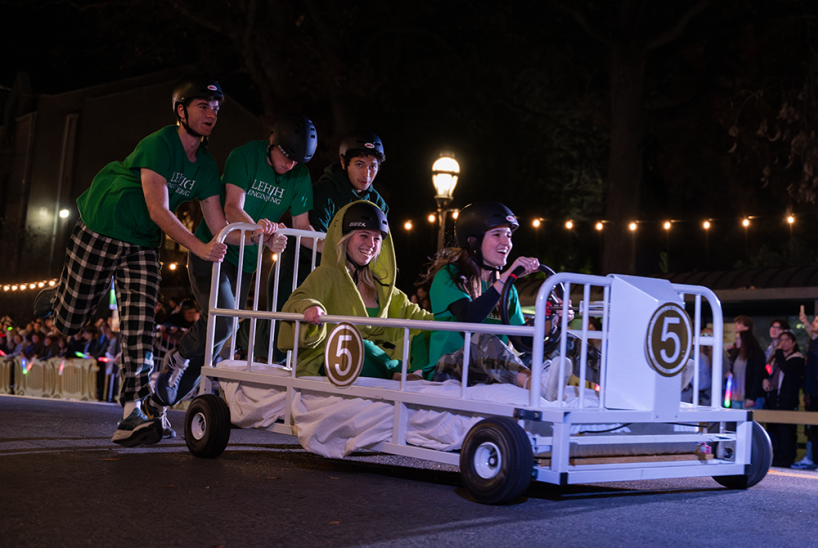 Lehigh students dressed in green Lehigh Engineering shirts race bed-shaped cars down a road.