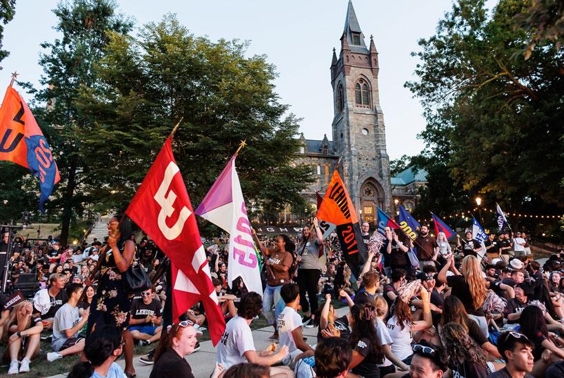 A large group of people sits and holds colorful class flags on a grassy area in front of the Clayton University Center at Lehigh University.