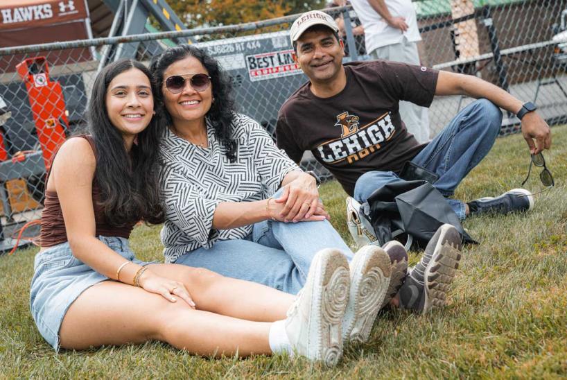 A family of three smiles while sitting together on the grass at an outdoor Lehigh University event near a chain-link fence.