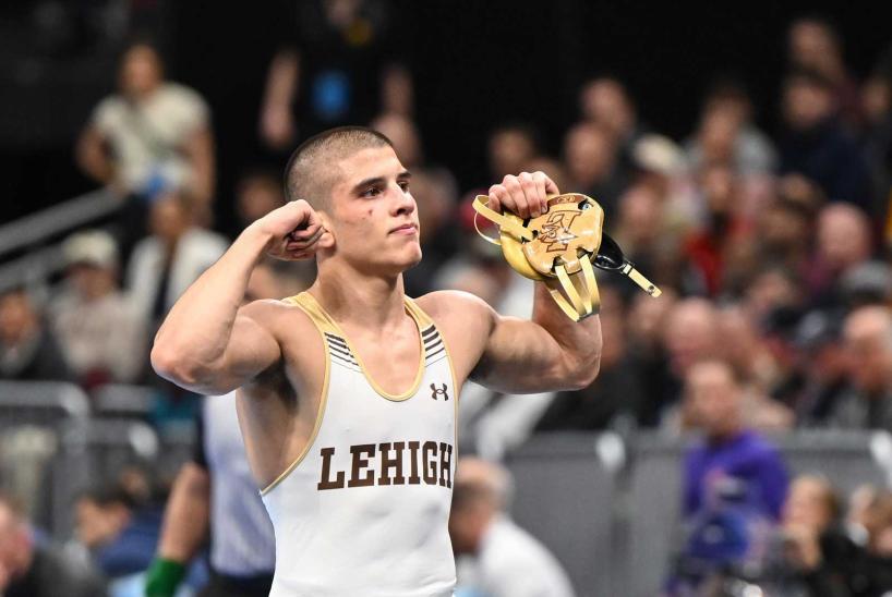 A Lehigh wrestler flexes his biceps while holding his brown and gold headguard to celebrate victory in front of a crowd.