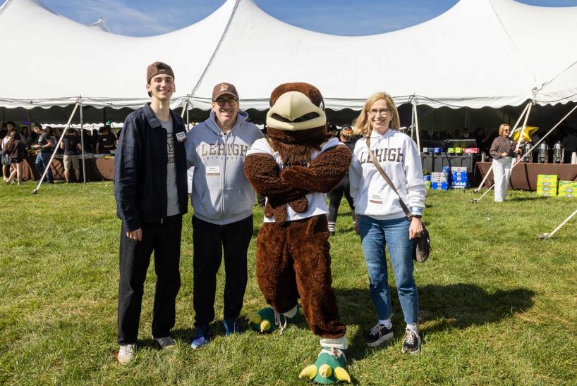 Lehigh University mascot Clutch stands with a family of three on a grassy field in front of a large white event tent during Lehigh Family and Founder's Weekend.