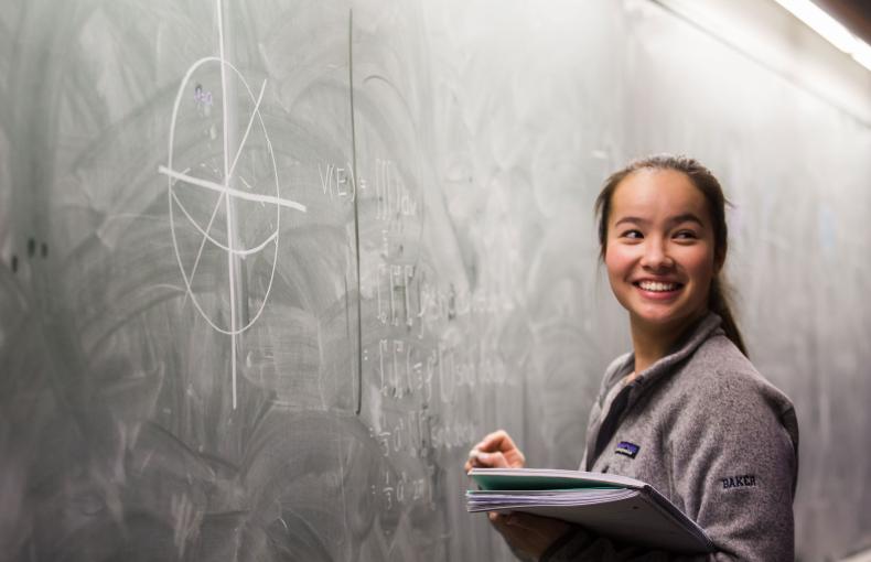 A student smiling at a blackboard holding her notebook