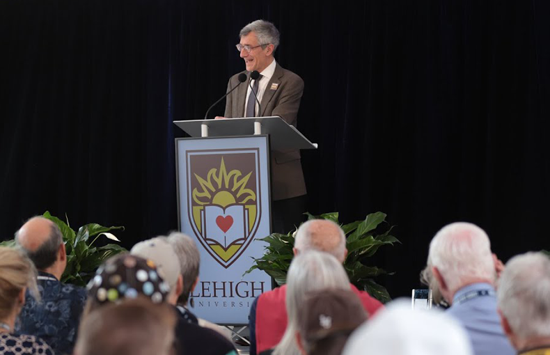 Lehigh President Joseph Helble stands at the podium designed with a large university crest with a crowd of event attendees.
