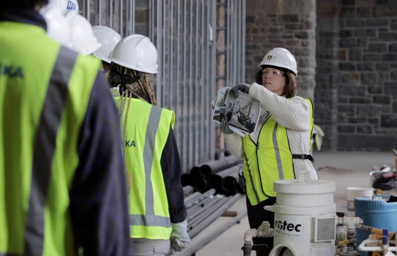 A woman leads a group through a construction site, all wearing white hardhats and yellow vests, and shows them a photo rendering.