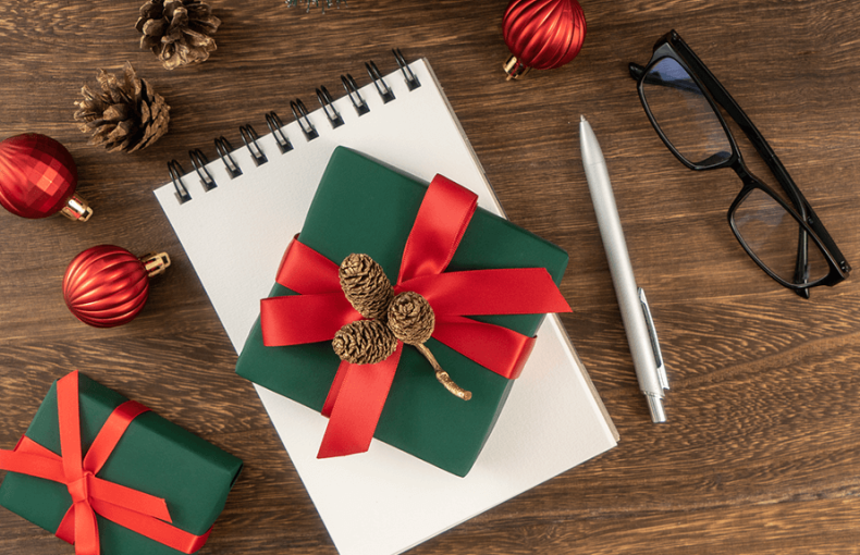 a small gift wrapped with ribbon and decorated with pinecones sit atop a notepad with a pen, glasses, and ornaments surrounding it