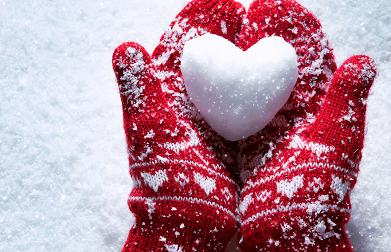 Hands wearing red wool mittens delicately hold a snowball shaped like a heart against the backdrop of a blanket of white shimmering snow.