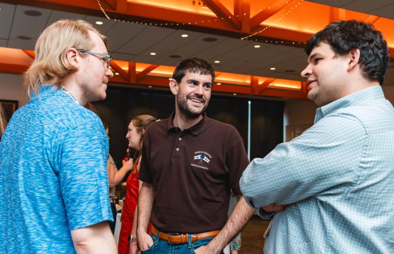 Three male Lehigh alumni in casual attire talking at an indoor reception with warm architectural lighting.
