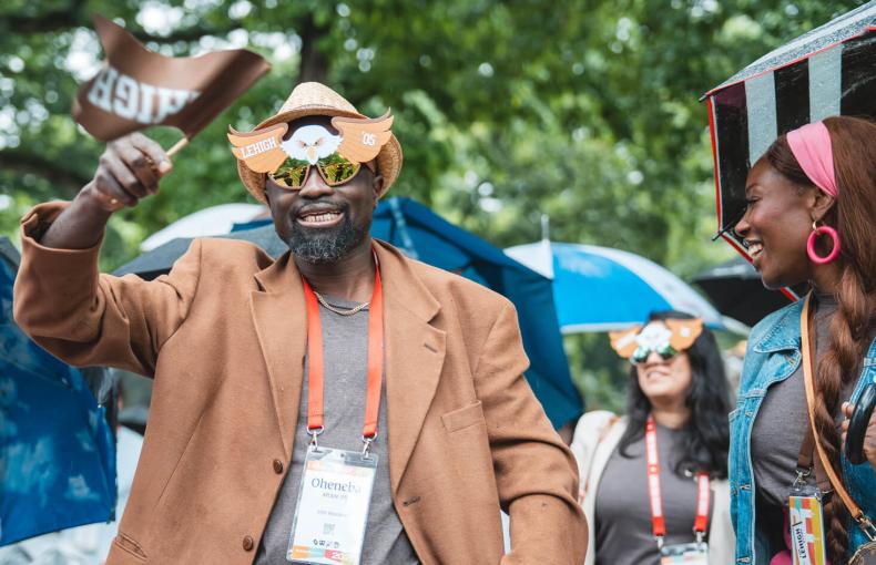 A Lehigh alumnus in eagle-themed sunglasses and a straw hat waving a school flag during a rainy outdoor parade.