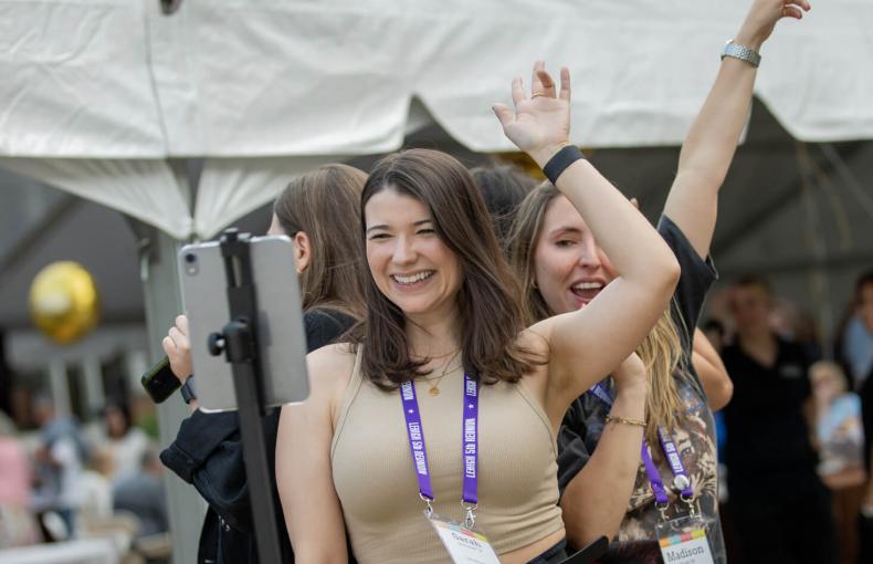 A small group of women laughing and dancing near a white event tent with string lights and a tablet on a tripod.