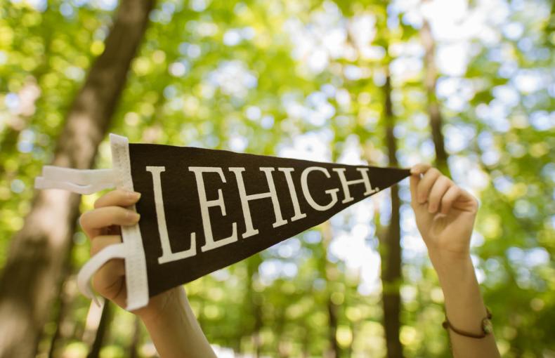 Hands hold up a brown and white Lehigh pennant against a blurred background of sunlit green trees.