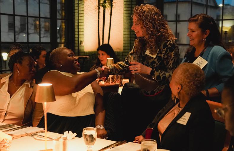 Two women shake hands across a dinner table during a Lehigh alumni event in a dimly lit, upscale restaurant.