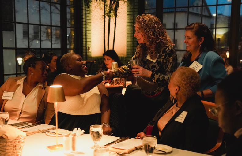 Two women shake hands across a dinner table during a Lehigh alumni event in a dimly lit, upscale restaurant.