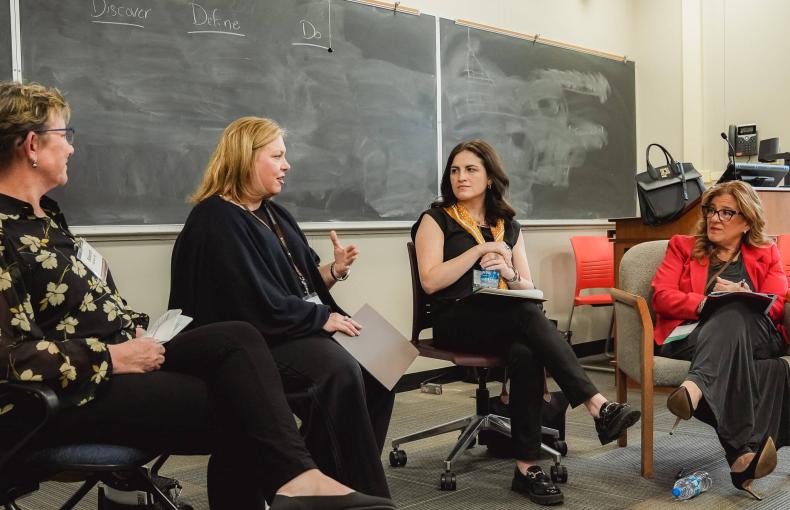 Four women participate in a panel discussion in a classroom, seated in front of a chalkboard with "Discover Define Do" written on it.