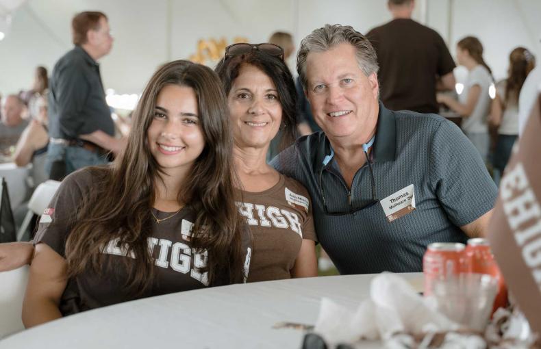 A family of three sit together at a white covered table, smiling.