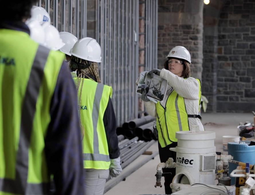 A woman leads a group through a construction site, all wearing white hardhats and yellow vests, and shows them a photo rendering.