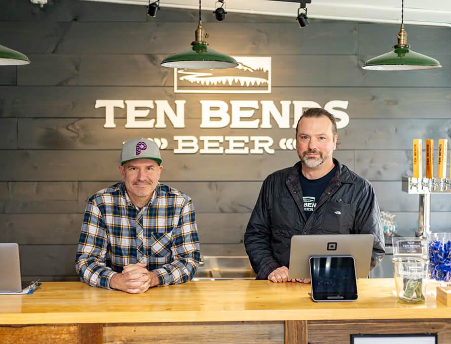 Mike Scarlata and Jason Powell stand at a light wood counter with the Ten Bends Beer logo on the wall behind them
