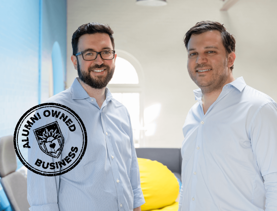 In a brightly lit room with baby blue walls, two men stand casually facing each other and smiling for the camera and an "Alumni-owned business" graphic overlays the photo.