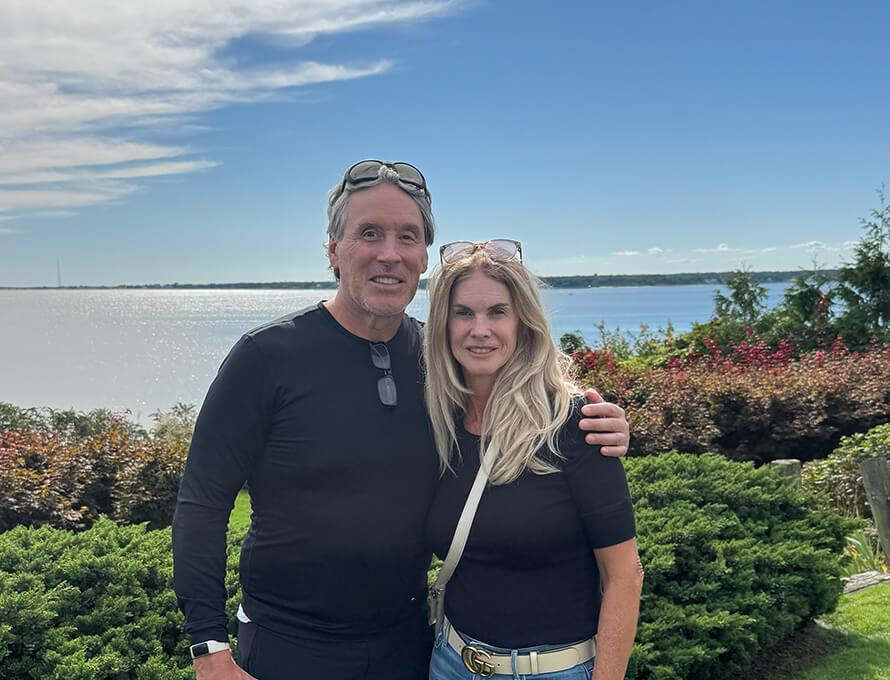A couple with their arms around each other smile on a sunny day by a scenic ocean view.