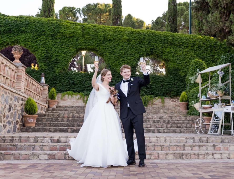 Eva and JJ hold up glasses of champagne on their wedding day; she wears a gown and holds their yorkie, he wears a black suit.