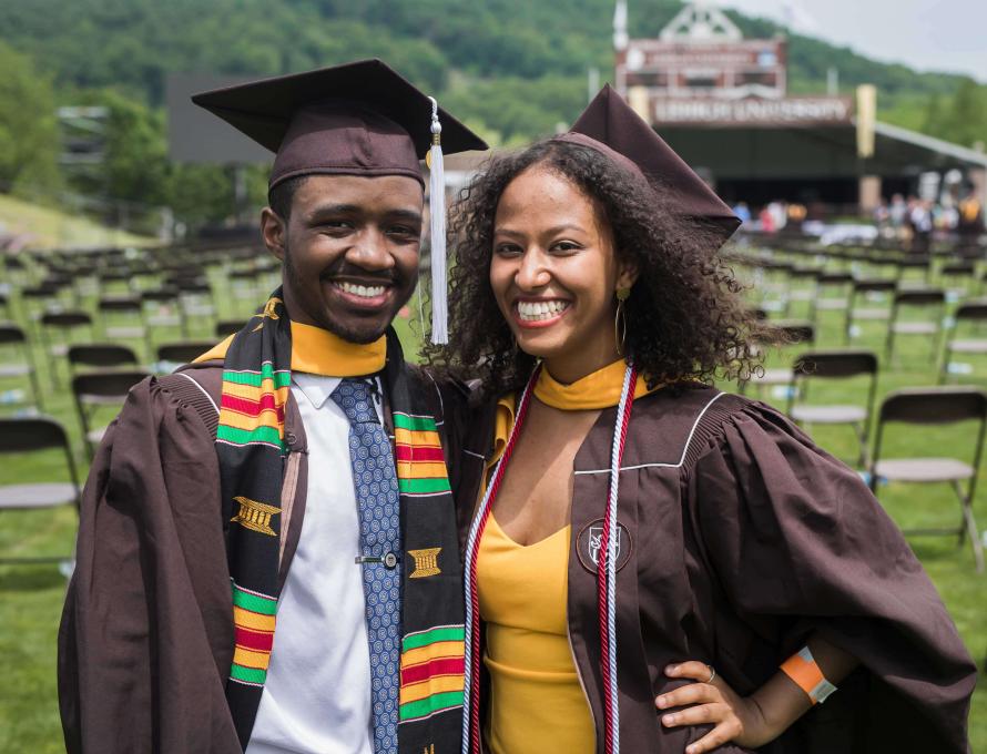 Jones and Taye stand and smile at graduation. Both wear brown caps and gowns, as well as gold hoods around their necks.