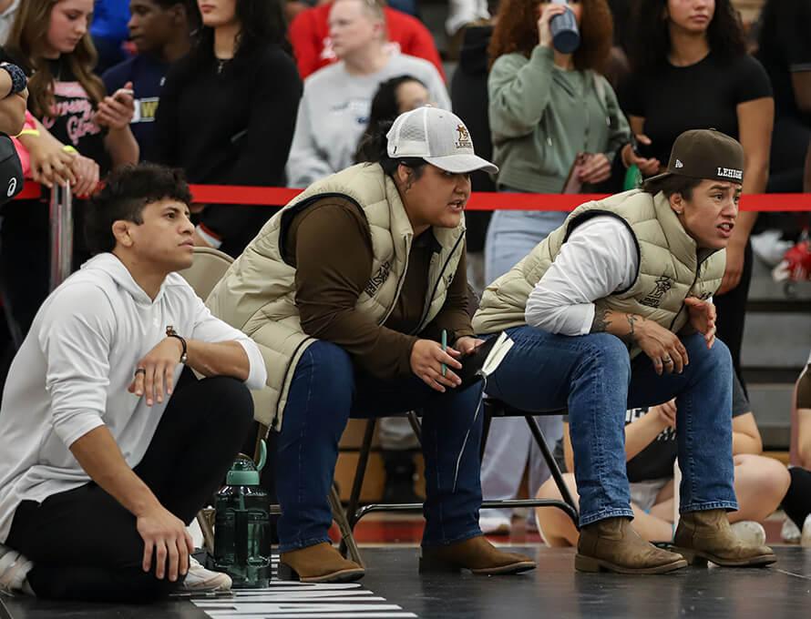 Three Lehigh wrestling coaches crouch at the edge of a gym mat, watching a match intently, while a crowd of spectators stands behind them.