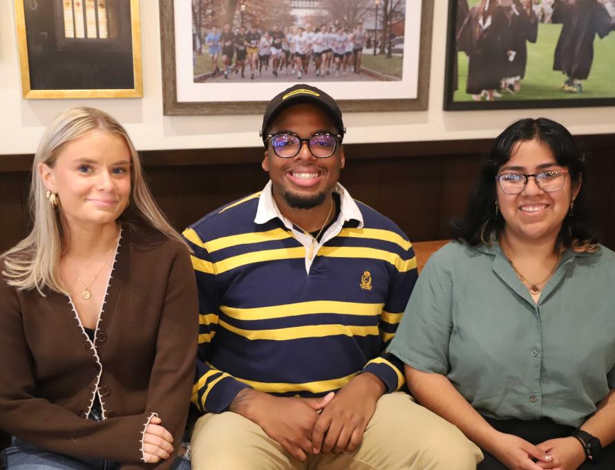 Caitlyn, Luke, and Crystal sit beside each other on a brown leather couch; photographs of students are behind them.