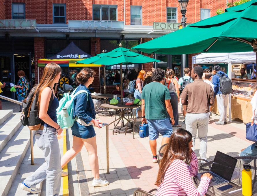 People walk around the Farmers' Market on a sunny day, many of them wearing backpacks. Bright green umbrellas shade tables.