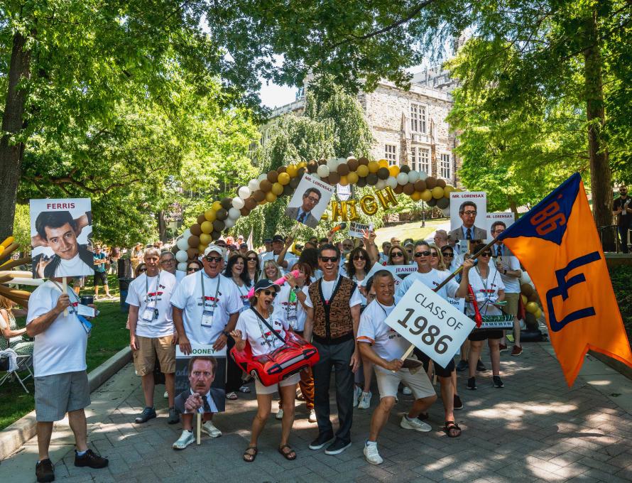 Members of the Class of 1986 smile and hold up signs with characters from Ferris Bueller's Day Off as they wave their class flag. 
