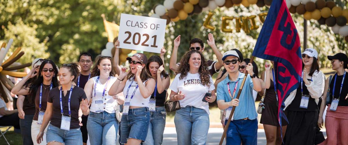 A cheerful group marches outdoors during a Lehigh University reunion, holding a "Class of 2021" sign and carrying a university flag under a balloon arch.