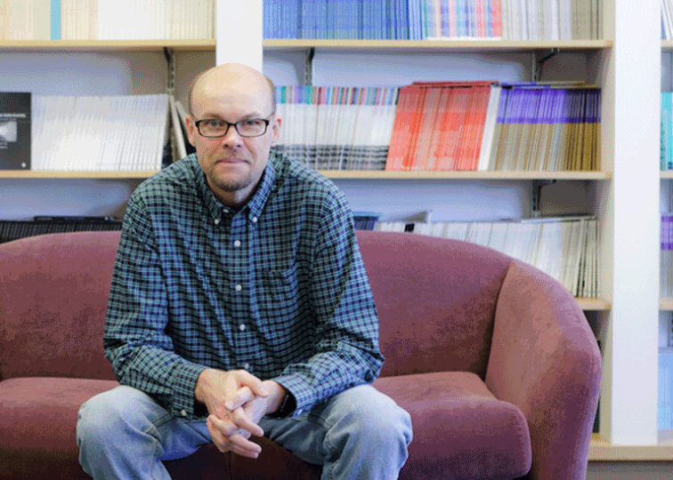 Jeremy Littau sits on a couch with shelves of books behind him