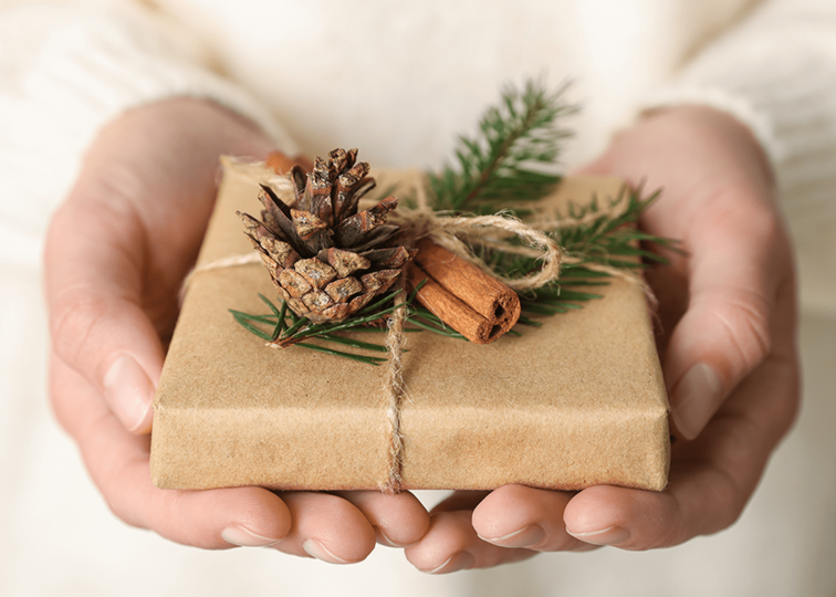 A pair of hands holding a holiday present wrapped in brown paper, topped with a pine cone and cinnamon stick, and bound by twine.