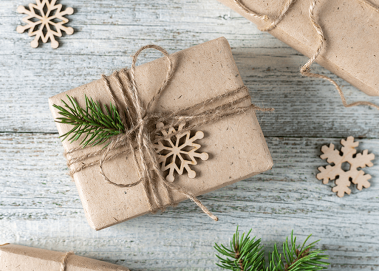 High angle view of brown paper wrapped gift, tied with natural twine, a sprig of a pine branch, and small wooden snowflake ornament.