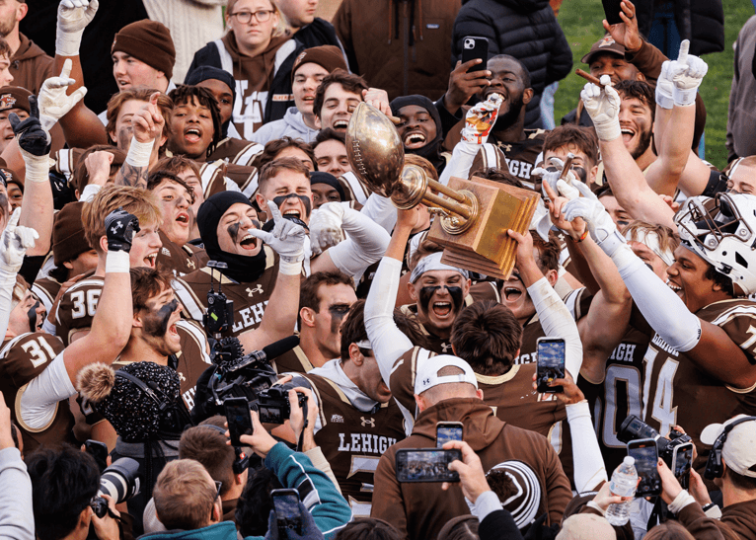 A crowd of Lehigh football players hold up a trophy and celebrate on the field of Goodman Stadium.