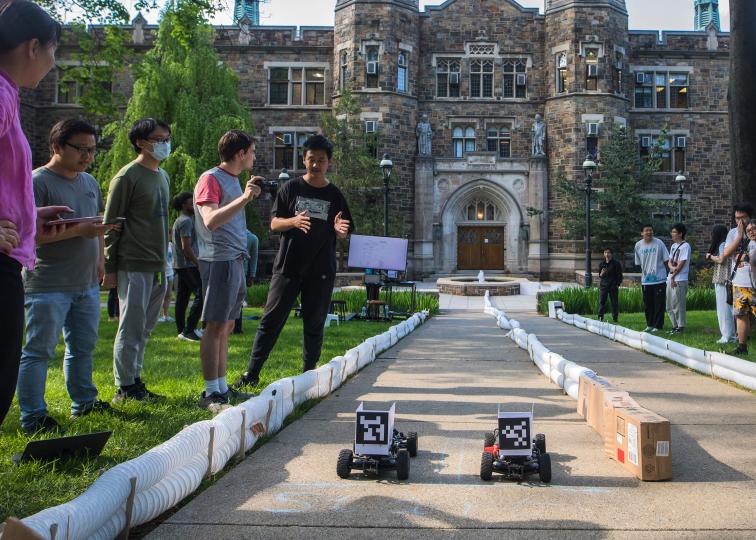 A race track is set up on the pathway in front of Packard Lab with small race cars competing   as competitors and spectators watch on from either side.