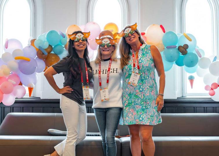 Three women wearing fun masks pose together in front of a balloon arch with pastel-colored balloons and a grey couch.