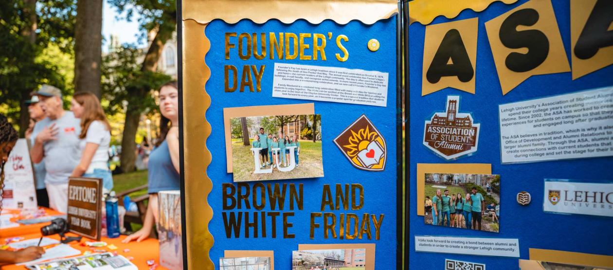 A blue tri-fold display board for the Association of Student Alumni featuring yellow lettering for campus traditions, student photos, and event info at an outdoor club expo.